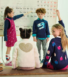 Four children in a classroom setting, each wearing a colorful sweater with educational graphics like letters, numbers, and apple motifs, standing and sitting in front of a periodic table board.