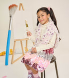 A young girl with dark hair in a ponytail sits on a stool, holding a paintbrush. She is wearing a white turtleneck sweater, a fuzzy lavender and pink sequined vest, and a pink and silver sequined skirt.