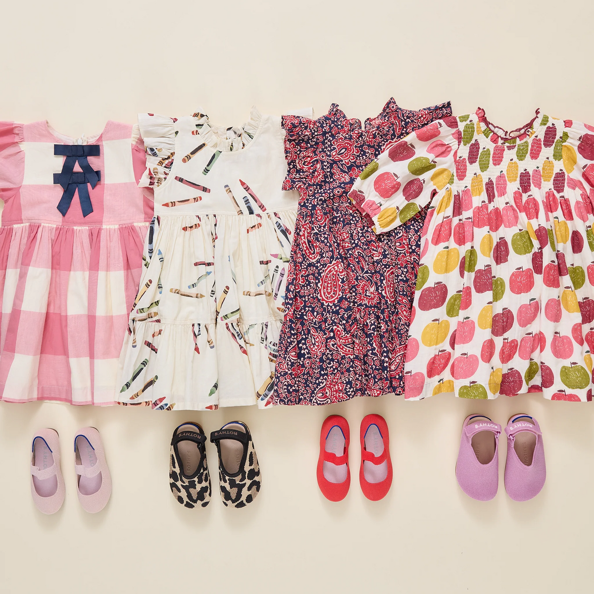 Four children's dresses are laid out side by side on a light beige background, each paired with a matching pair of flats. The dresses include pink and white gingham, white with a colorful print, a dark floral pattern, and a white print with colorful fruit motifs.