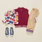 A styled flat-lay of a children's outfit set on a warm cream background, featuring a burgundy zip-up vest, a white long-sleeve shirt with a colorful apple print, beige pants with striped cuffs, navy blue canvas sneakers, and a small pencil.