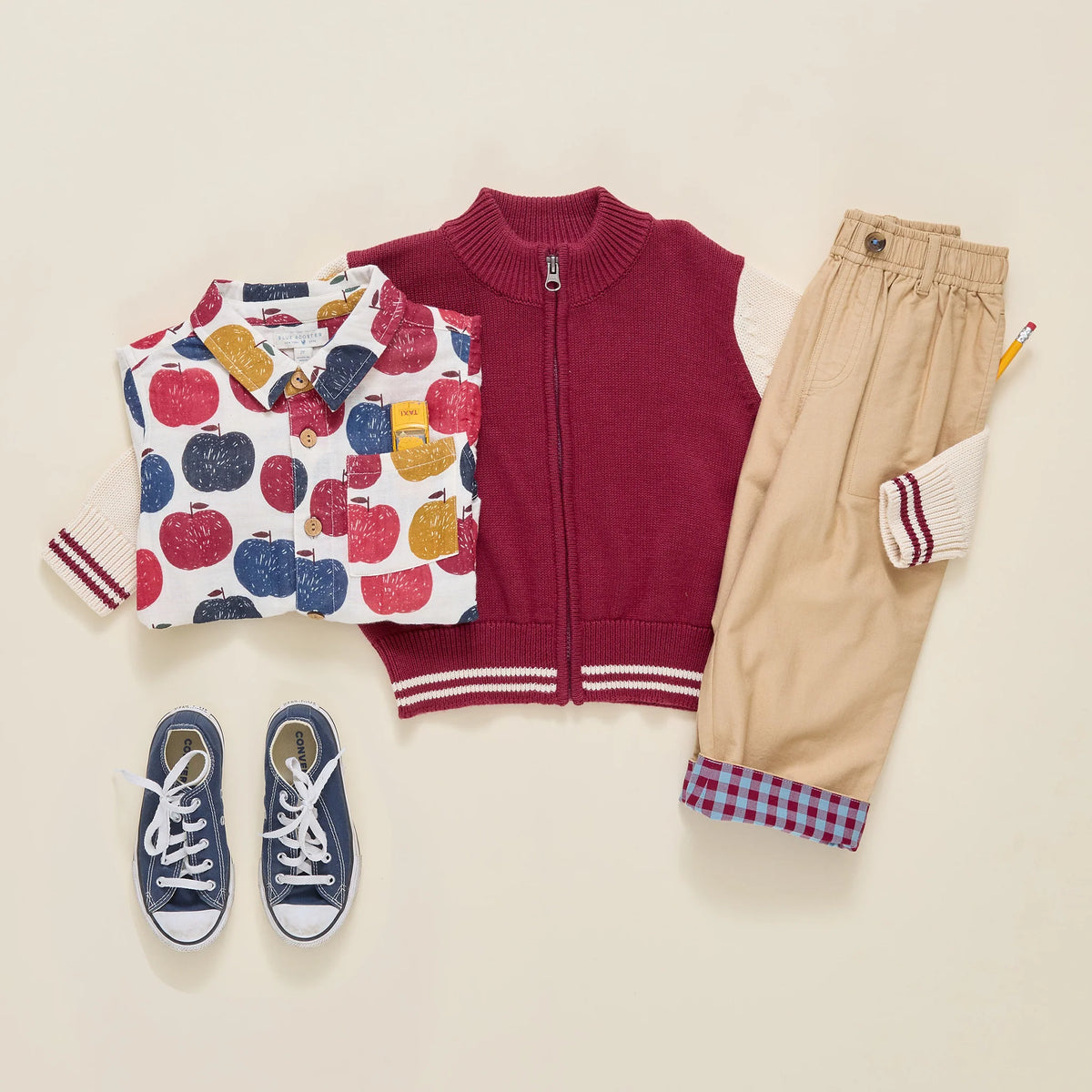 A styled flat-lay of a children's outfit set on a warm cream background, featuring a burgundy zip-up vest, a white long-sleeve shirt with a colorful apple print, beige pants with striped cuffs, navy blue canvas sneakers, and a small pencil.