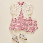 A baby girl's three-piece outfit set featuring a pink dress with a dog print, a cream eyelet cardigan, and a matching pink ruffled skirt, styled with a pair of silver baby shoes on a cream background.