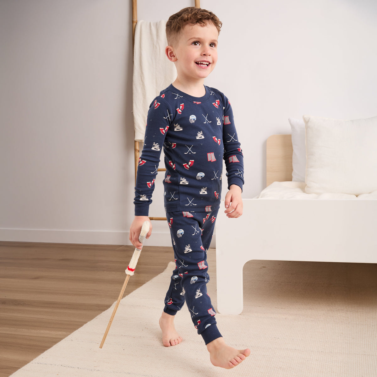 A young boy wearing a navy blue long-sleeve pajama set with a pirate-themed print, walking barefoot on a light-colored rug in a bright bedroom.