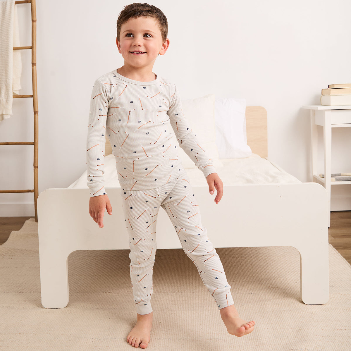 A young boy stands barefoot in front of a white bed wearing a long-sleeve pajama set with a small scattered print of orange pencils, blue dots, and brown lines on an off-white background.