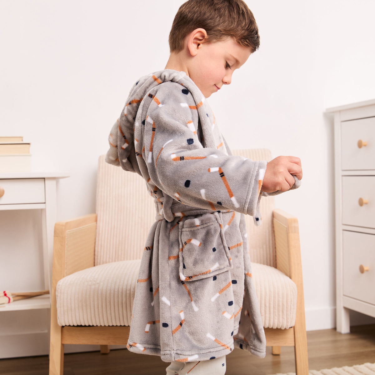 A young boy wearing a gray plush hooded bathrobe with a pattern of orange, white, and navy pencils, standing in a child's bedroom next to a light wood armchair.