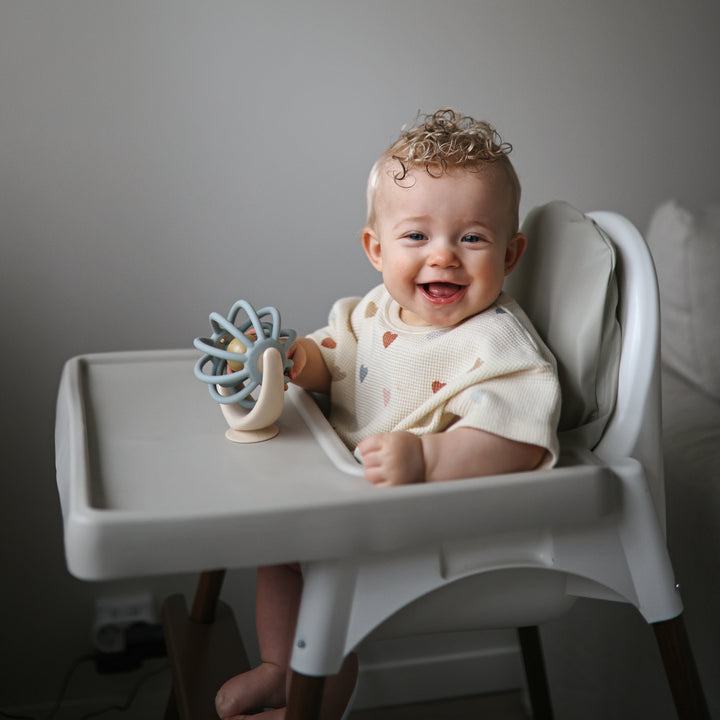A smiling baby sitting in a white high chair, looking at and touching a blue and beige Mushie Suction Spinner Ball attached to the tray.