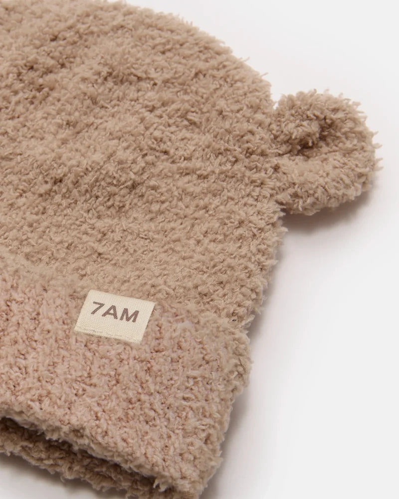 A close-up of a beige sherpa beanie with a small pom-pom on one side and a folded cuff featuring a cream-colored '7AM' brand label.