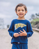 A young boy wearing a navy blue sweatshirt and matching sweatpants stands outdoors. The sweatshirt features a yellow and white 'LET'S GO!' graphic on the front.