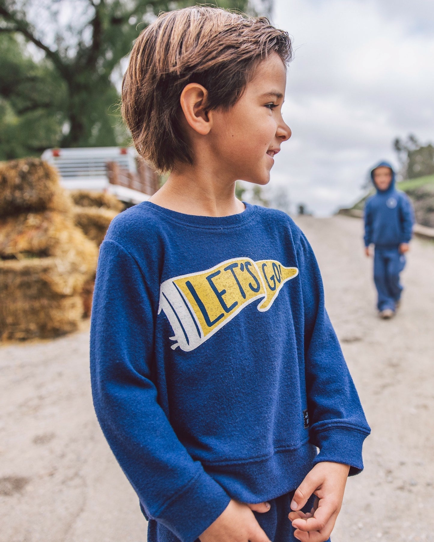 A young boy wearing a royal blue crewneck sweatshirt with a yellow and white 'LET'S GO' pennant graphic, standing outdoors on a dirt path with hay bales in the background.