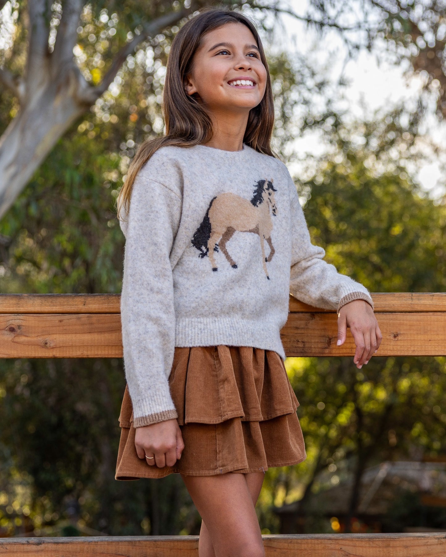 A young girl smiling and looking up while wearing a light gray knit sweater with a brown and beige horse design on the front, paired with a brown tiered skirt, leaning on a wooden fence outdoors.