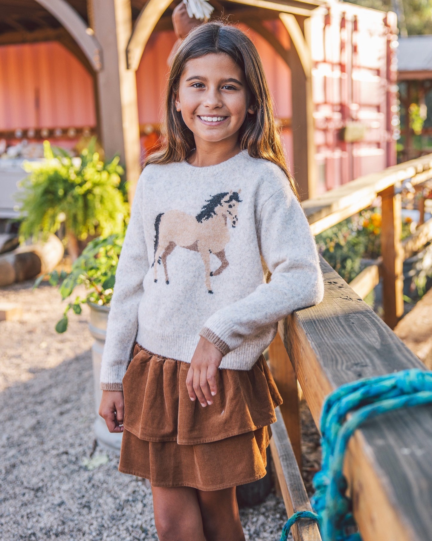A young girl smiling while wearing a light gray knit sweater with a horse graphic and a brown tiered skirt, standing outdoors near a wooden railing.