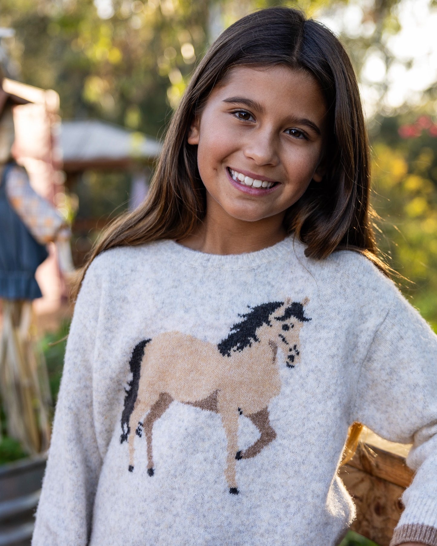 A young girl smiling while wearing a light gray knit sweater with a large horse graphic in tan, brown, and black on the front, photographed outdoors with a blurred background.