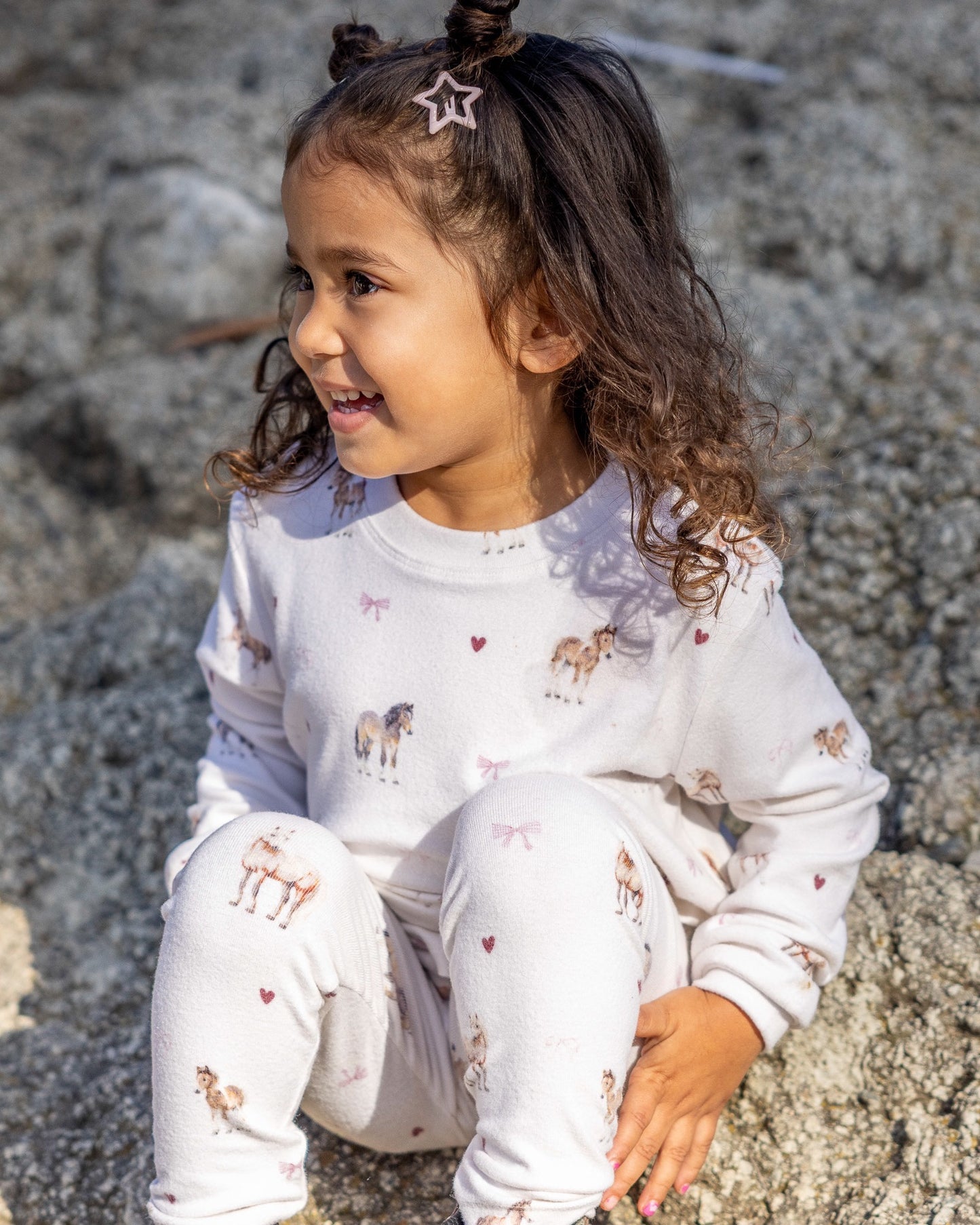 A young girl with curly brown hair sits on a rock outdoors, wearing a white long-sleeve pajama set with a cute print of donkeys and small pink hearts.