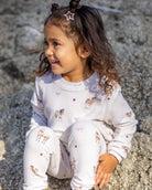 A young girl with curly brown hair sits on a rock outdoors, wearing a white long-sleeve pajama set with a cute print of donkeys and small pink hearts.
