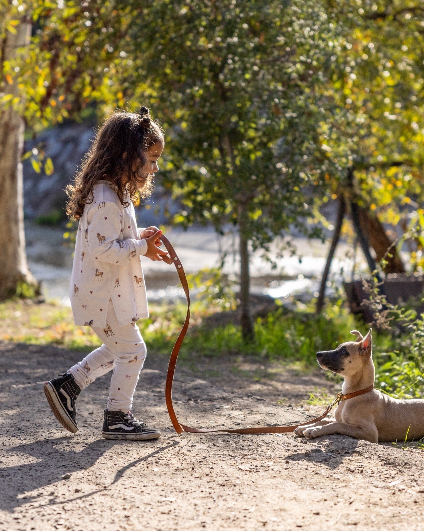 A young girl in a white patterned long-sleeve top and matching pants walks outdoors on a dirt path while holding a brown leash attached to a tan dog lying on the ground. Trees and foliage are visible in the background.