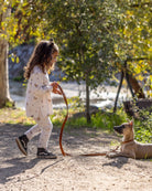 A young girl in a white patterned long-sleeve top and matching pants walks outdoors on a dirt path while holding a brown leash attached to a tan dog lying on the ground. Trees and foliage are visible in the background.