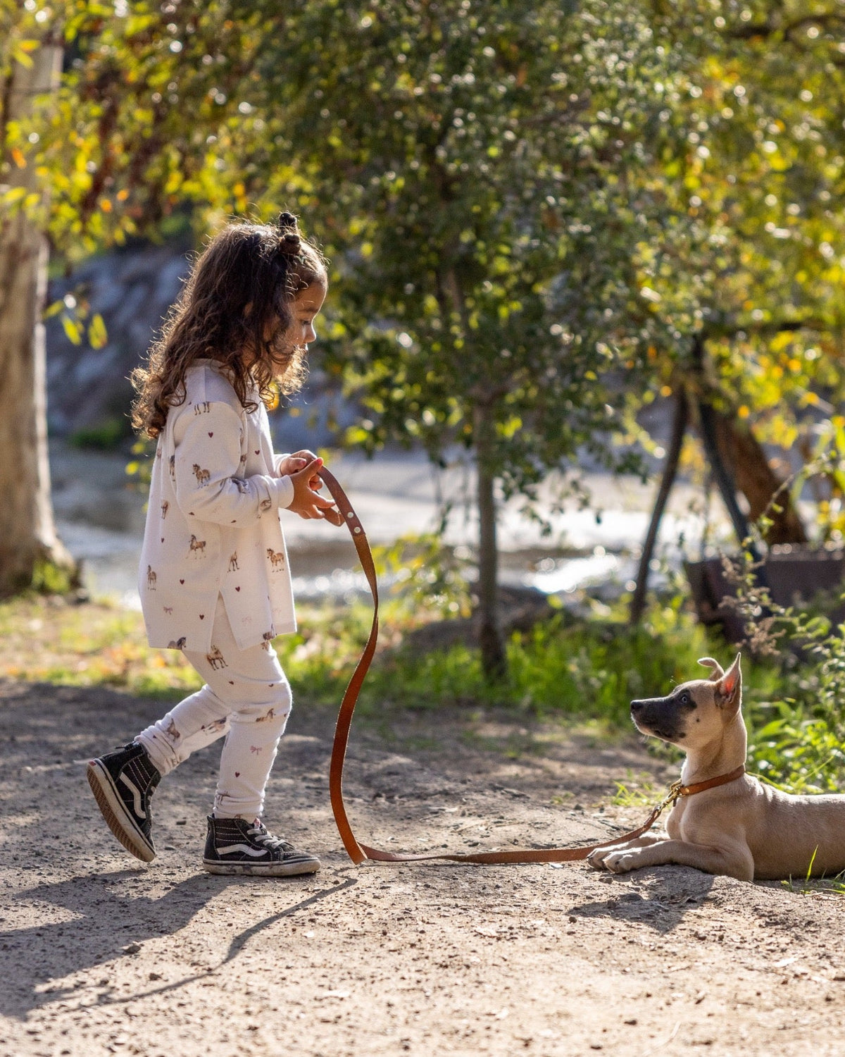 A young girl in a white patterned long-sleeve top and matching pants walks outdoors on a dirt path while holding a brown leash attached to a tan dog lying on the ground. Trees and foliage are visible in the background.