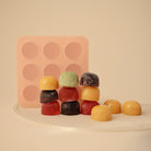A pink silicone mold stands upright next to a colorful stack of various frozen baby food cubes and individual portions arranged on a flat surface.