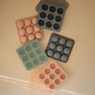 An assortment of silicone baby food freezer trays and lids in various colors, including green, peach, and grey, arranged on a flat surface.