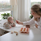 A mother smiling while feeding baby food with a spoon to a baby sitting in a high chair, with a silicone baby food tray on the table in front of them.