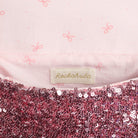 Close-up of a children's garment showing a light pink fabric with a subtle bow print, a central cream fabric label reading Rockahula, and a sequined hem in pink and burgundy sequins.