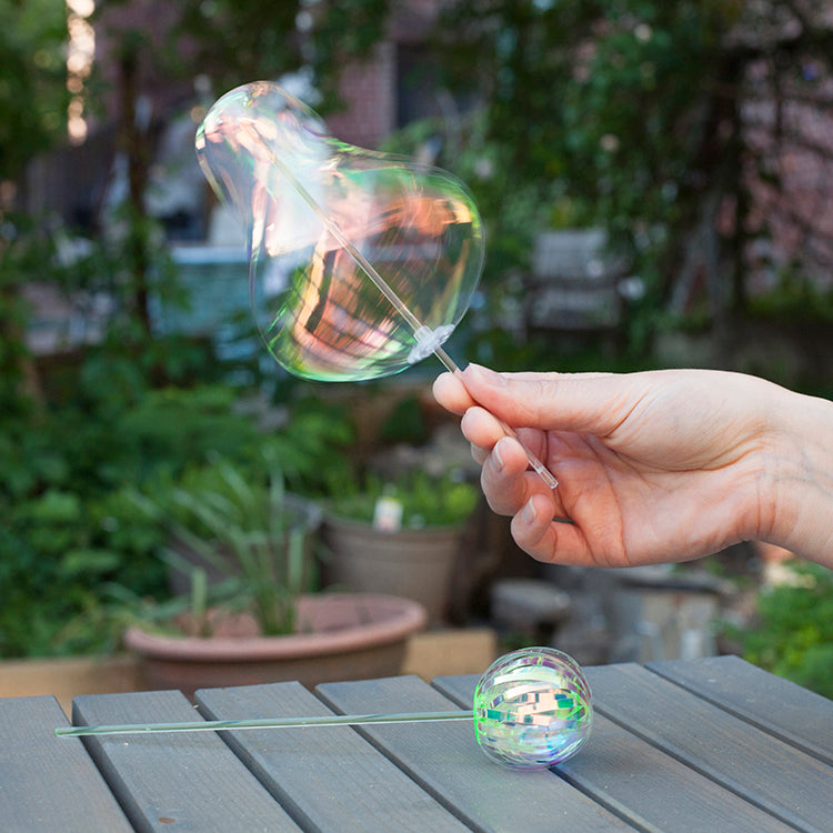 Hand holding a bubble wand with a bubble, set against a garden background