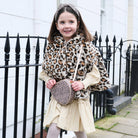 A young girl wearing a leopard-print faux fur hooded cape over a cream dress, holding a matching leopard-print crossbody bag and wearing a leopard-print headband, standing outdoors in front of a white building and black metal fence.