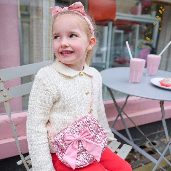 Young girl with a pink bow in her hair, wearing a white sweater and red pants, sitting on a bench with a pink sparkly bag.