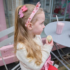 Young girl with a pink headband holding a donut, sitting at a table with a pink drink.