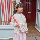A young girl wearing a cream cable-knit cardigan, pink skirt, and a gold floral headband carries a heart-shaped crossbody bag with a red cherry pattern and red strap, standing in front of a red door.