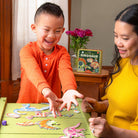 A child and an adult playing the Dinosaur Dice Game by Eeboo together at a table, with colorful dinosaur fossil puzzle pieces spread out and the game box visible in the background.