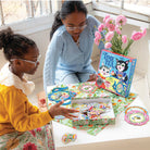 Two young girls sitting at a table indoors, happily playing with the Kitten's Time for Tea Spinner Game by Eeboo, with game pieces, a box, and a vase of pink flowers nearby.