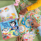 Kitten's Time for Tea Spinner Game by eeBoo box and game components laid out on a floral tablecloth, with a child spinning the teapot spinner and colorful cutouts of tea party treats nearby.