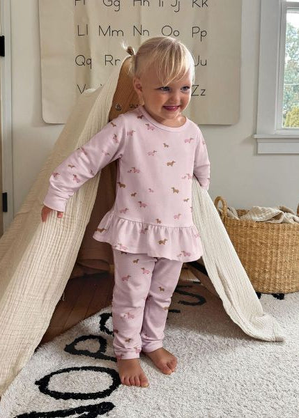 A young child wearing a light pink ruffled two-piece sweatsuit with an all-over dog print by günamüna.