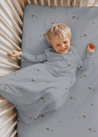 A smiling toddler lies on their back in a wooden crib, wearing a long-sleeve sleep sack and matching fitted crib sheet. The fabric is a dusty slate blue with a small brown and white deer print, set against a neutral cream-colored crib.