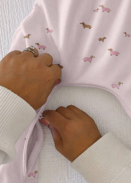 A close-up of a baby garment being fastened by hands, showing a pale pink fabric with a small dog print and a white ribbed sleeve.