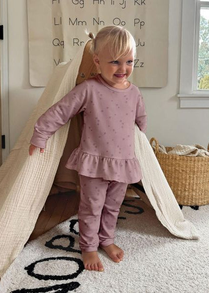 A toddler stands barefoot inside a fabric teepee play tent in a bright nursery. The child is wearing a dusty mauve long-sleeve top and matching pants with a subtle stars pattern.
