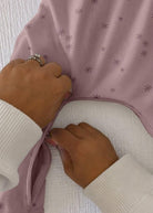 A close-up of a dusty pink baby garment with a small star pattern, being held open by two hands wearing white ribbed long-sleeve shirts.