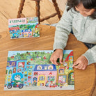 A child sitting at a wooden table working on the 20 Piece Puzzle - Yumi's Village by Eeboo, with the colorful puzzle box resting on the table above the completed village scene.