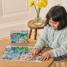 A child sitting at a wooden table working on the 20 Piece Puzzle - Yumi's Village by Eeboo, with the colorful puzzle box resting on the table above the completed village scene.