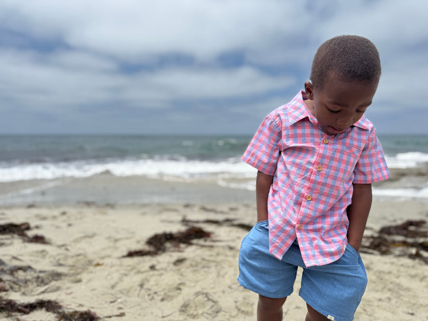 Child in a checkered shirt and shorts standing on a beach with ocean waves in the background