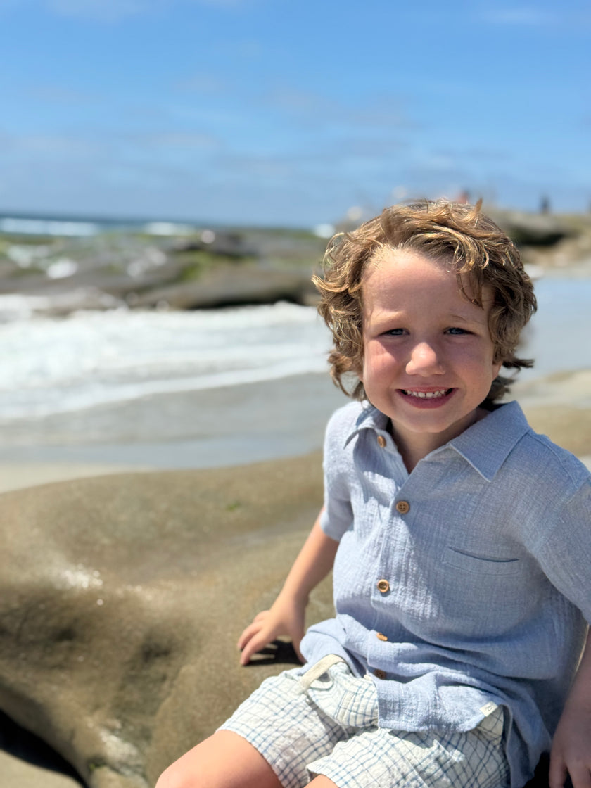 Child sitting on a rock by the ocean with a clear blue sky.