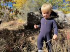 A toddler with blonde hair wearing a navy blue Henley onesie stands outdoors in a natural setting with rocks and dry brush, holding twigs.