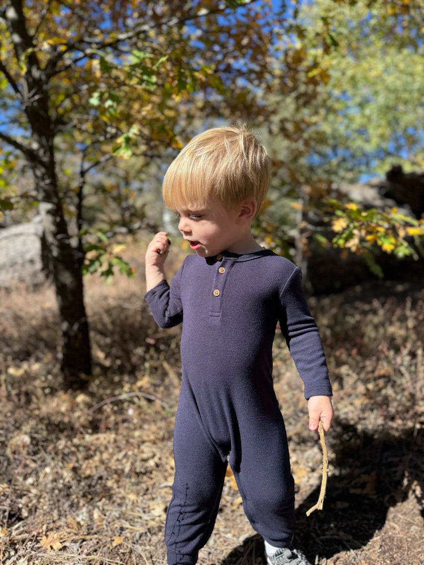 A toddler with blonde hair wearing a dark navy long-sleeve footless onesie with a three-button henley collar, standing outdoors in a wooded area with autumn trees.