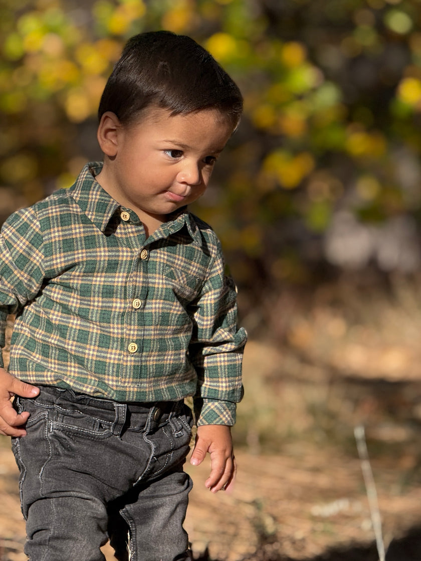 A toddler boy wearing a green and beige plaid long-sleeve button-up shirt and dark gray jeans, standing outdoors in a sunny, blurred natural setting.