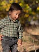 A toddler boy wearing a green and beige plaid long-sleeve button-up shirt and dark gray jeans, standing outdoors in a sunny, blurred natural setting.