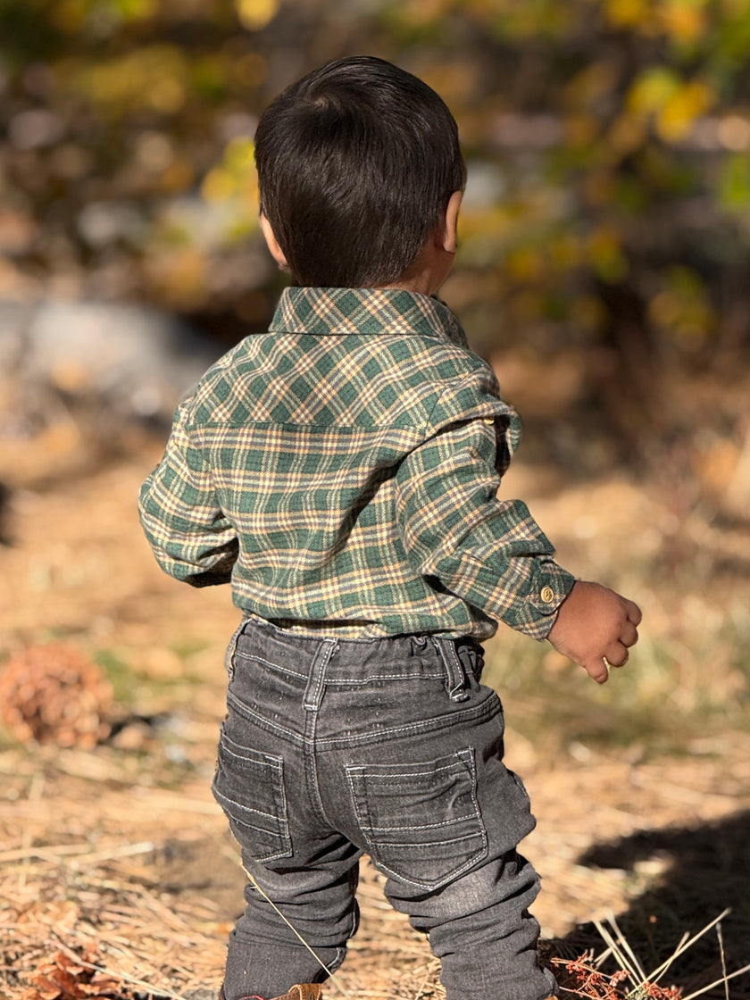 A toddler viewed from behind wearing a green and beige plaid long-sleeve button-up shirt and dark gray jeans, standing outdoors in a sunny, dry grassy area with blurred trees in the background.