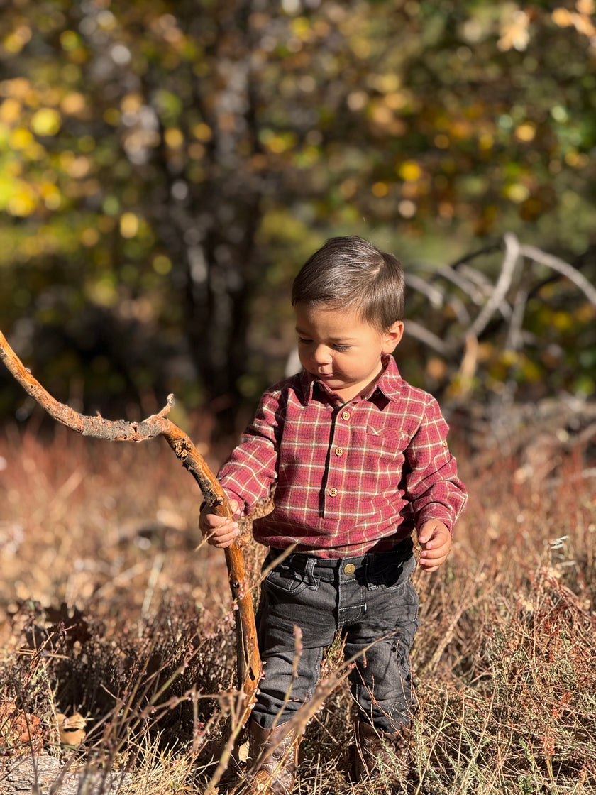 Child holding a stick in a natural setting with trees and dry grass, wearing a red and green plaid button-down shirt and dark pants.