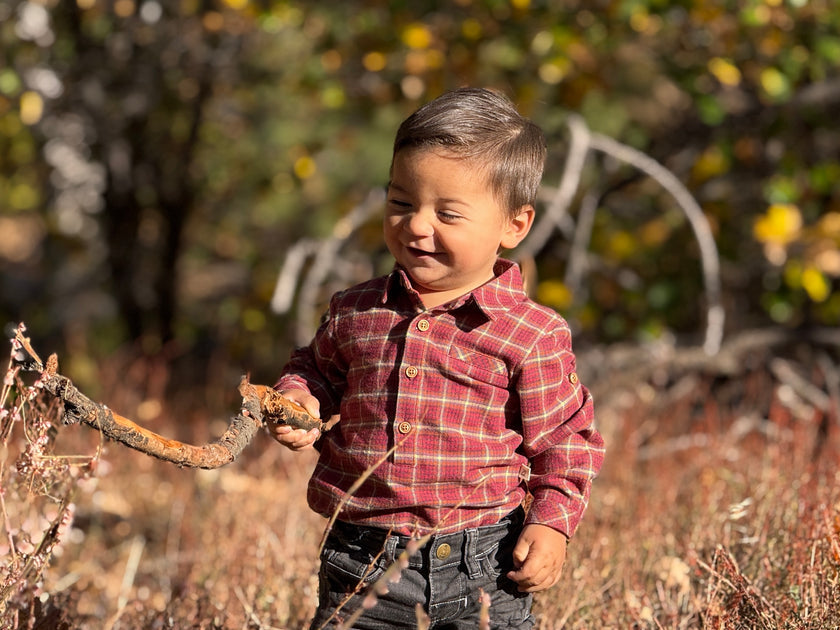 A young boy wearing a red and yellow plaid button-up shirt and dark jeans stands outdoors in a grassy field, holding a small branch.