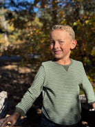 Young boy standing in a forested area with trees and dry grass in the background.